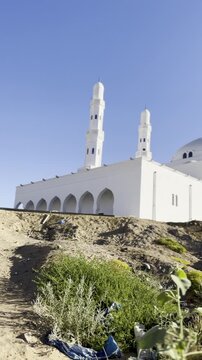 A scenic view of Masjid Al-Areesh featuring its iconic white minarets, dome, and traditional mashrabiya windows against a rugged desert landscape and clear blue sky.