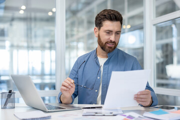 Professional man in smart-casual attire reviewing business documents at a bright modern desk, holding glasses and smiling confidently while working on strategy and finances by laptop