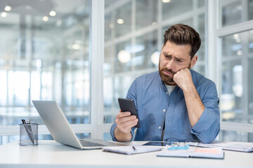 Bored businessman feeling unproductive and procrastinating, sitting at office desk checking smartphone while laptop and work documents remain untouched, representing job burnout and distraction
