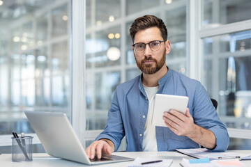 Professional man with glasses and beard working at desk in a bright modern office, using laptop and holding tablet while analyzing data and planning strategy, looking confident and focused