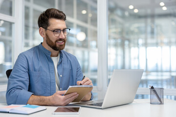 Positive bearded businessman working on a laptop, smiling and writing in a notebook while participating in a virtual conference or remote work session in a modern office