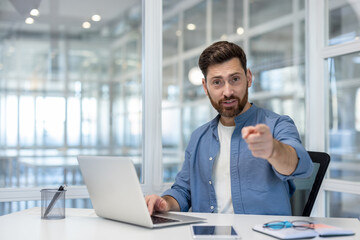 Confident young businessman on a laptop video call in a modern office, pointing at the camera while leading a virtual meeting or presentation, engaged and assertive