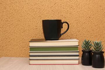 Black mug on top of a pile of books, on a wooden table, in front of orange color wall. Horizontal shot with copy space, perfect for study themes, course coffee break, and education contents.
