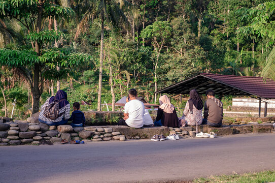 A group of people sit by a roadside in a rural area, near trees and fields, during the day, resting together as they socialize and enjoy a calm countryside moment.