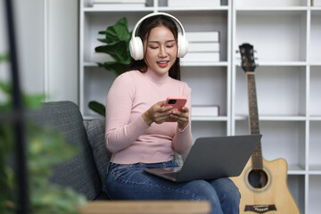 Happy young woman sitting on comfortable sofa, wearing headphones and using laptop computer and smartphone while listening to music in living room at home.