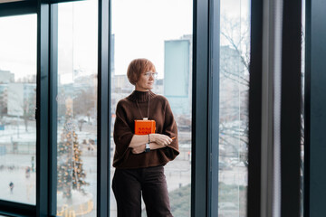 Confident professional woman standing by window holding planner