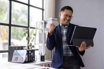 Young entrepreneur feeling euphoric while looking at tablet, celebrating successful business deal or good news in modern office.