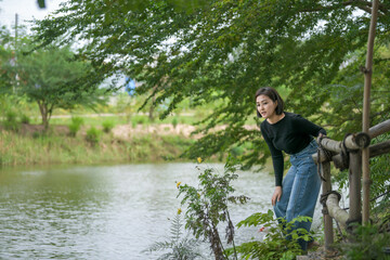 A woman in a black top and jeans stands at the edge of a pond, watching three white ducks swimming peacefully in the water surrounded by nature.