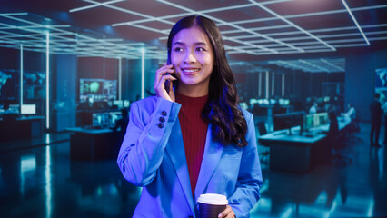 Smiling Asian professional woman in blue blazer uses mobile phone and holds coffee cup with futuristic IT room computer monitor office featuring neon lights and modern digital technology background.