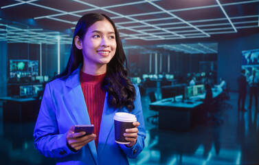 Smiling Asian professional woman in blue blazer uses mobile phone and holds coffee cup with futuristic IT room computer monitor office featuring neon lights and modern digital technology background.