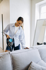 Woman vacuuming sofa at home using rechargeable vacuum cleaner