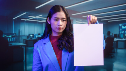 Smiling Asian woman holds blank white paper in high-tech office. This mockup template offers perfect copy space for business messages, advertisements, or announcements in futuristic setting.