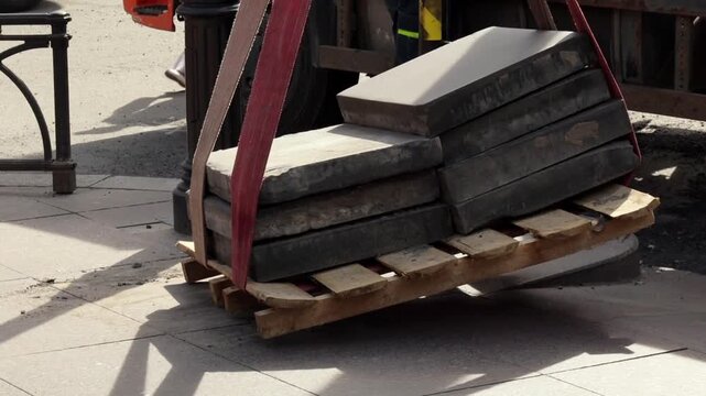 Stack of paving slabs sits on wooden pallet secured with lifting straps near truck wheel. Sidewalk paving and construction setting visible.