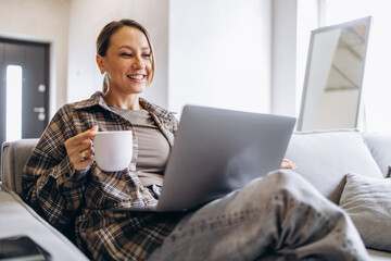 Woman working online from home, using laptop sitting on sofa and drinking coffee