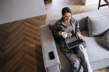 Woman working online from home, using laptop sitting on sofa and drinking coffee