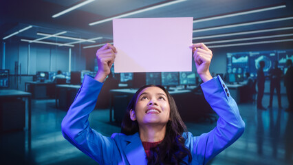 Smiling Asian woman holds blank white paper in high-tech office. This mockup template offers perfect copy space for business messages, advertisements, or announcements in futuristic setting.