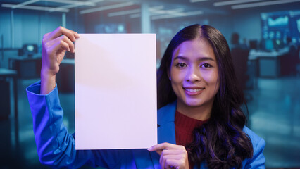 Smiling Asian woman holds blank white paper in high-tech office. This mockup template offers perfect copy space for business messages, advertisements, or announcements in futuristic setting.