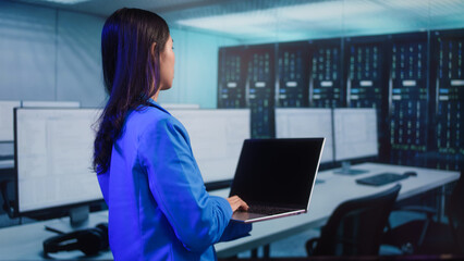 Serious Asian woman works on laptop while standing in high-tech workspace. digital tasks, data analysis, remote corporate connectivity, technology usage, data analysis, productivity, innovation