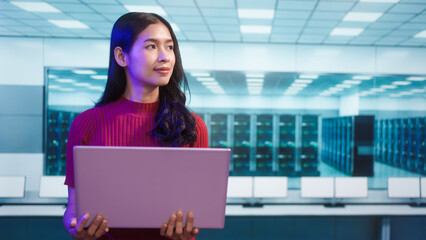 Focused Asian female IT professional works on laptop in secure data center. The modern background features server racks, cybersecurity, cloud computing, advanced digital infrastructure management.