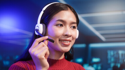Friendly Asian customer support call center agent wears headset in high-tech control room. She provides professional online assistance and manages communications in busy digital operations center.