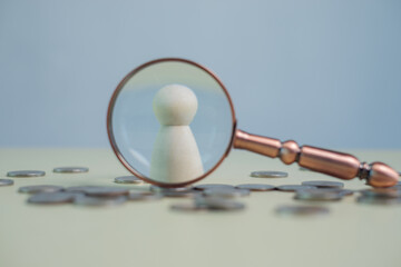 Wooden figure under magnifying glass with coins on table, representing financial analysis, personal finance review, money management, risk assessment, investment decision and economic inspection.