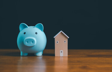 Blue piggy bank next to wooden house model on table, symbolizing saving money for home, real estate investment, housing finance, mortgage planning, financial stability and future security.