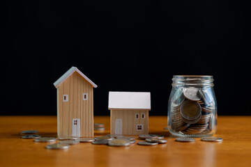Wooden house models with scattered coins and a glass jar of savings on wooden table, real estate finance concept for mortgage planning, property investment, home savings and wealth growth.