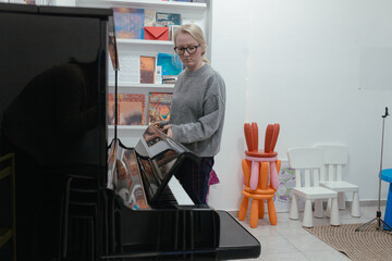 Student checks piano surface while standing inside classroom, reflective black finish shows surrounding shelves full music materials, posture signals active inspection, bright interior supports clear 