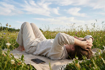 A young woman in linen clothing lies on her back with hands behind her head on a blanket in a grassy field of wildflowers, relaxing next to her mobile phone, symbolizing a digital detox and leisure.