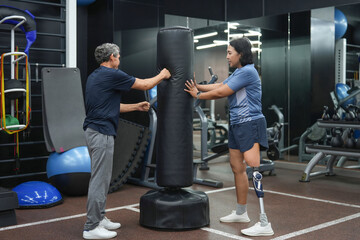 woman with prosthetic leg fitness trainer coach a senior man client boxing with free-standing punching bag at indoor gym
