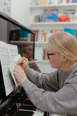 Music tutor writes pencil marks over sheet notes during piano lesson inside classroom with bookshelves full of learning materials © Kseniia