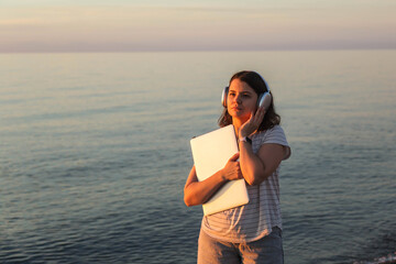 Woman holding laptop and listening to music on seaside at sunset, remote work and digital nomad lifestyle, wireless headphones, calm mood, freedom, focus and inspiration, copyspace