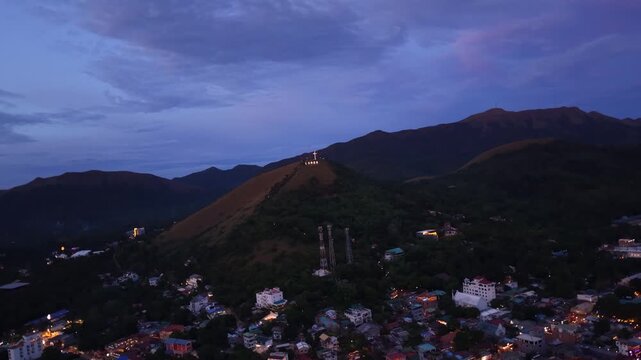 Cinematic rotating aerial footage reveals Mount Tapyas illuminated cross overlooking Coron town during blue hour. City lights twinkle below as twilight settles over Busuanga Island, Palawan