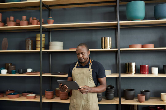 African potter using a digital tablet by shelves in a ceramics workshop