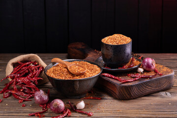 Chili powder in black bowl and wooden spoon with garlic, shallots, spicy spice mix. Asian food ingredients arranged on wooden table. Scattered chili powder background.