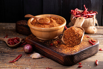 Chili powder in a wooden bowl and with a wooden spoon, along with garlic, shallots, and a mix of Asian spices, are arranged on a wooden table, conveying the concept of spicy food.