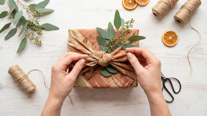 Top view of a Caucasian woman's hands tying a bow on a gift wrapped in earthy toned fabric using the Furoshiki technique. Eucalyptus sprigs, twine, and dried orange slices lay on a white wood.