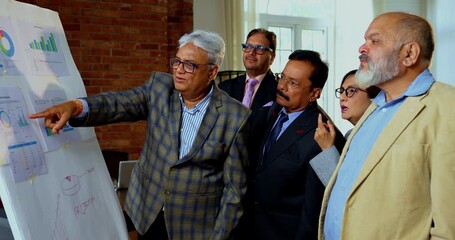 Indian business people discussing projection on chart while senior Asian executives stand close together reviewing growth data on white board inside modern corporate office meeting room - Powered by Adobe