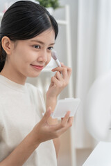 Beautiful Asian woman sit in front of a mirror and smile on makeup. face of a healthy woman applying makeup. Advertisement, lifestyle , cosmetics, makeup accessories, beauty activity, beautician