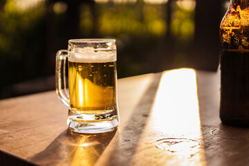 Golden beer in a clear glass mug rests on a wooden table, glowing in warm sunset light outdoor, refreshing beverage moment photography summer relaxation natural bokeh background.