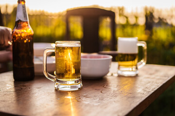 Golden beer in a clear glass mug rests on a wooden table, glowing in warm sunset light outdoor, refreshing beverage moment photography summer relaxation natural bokeh background.