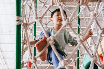 Active little Asian boy climbing on a rope net at an indoor playground. Child playing in a gym for physical development and muscle strength concept.