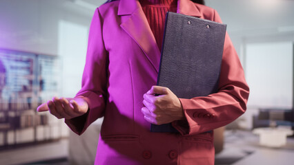 Close-up of a professional businesswoman in a pink blazer analyzing financial statistics on clipboard. She is reviewing data charts and writing notes in a bright, modern corporate office background.
