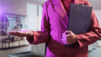Close-up of a professional businesswoman in a pink blazer analyzing financial statistics on clipboard. She is reviewing data charts and writing notes in a bright, modern corporate office background.