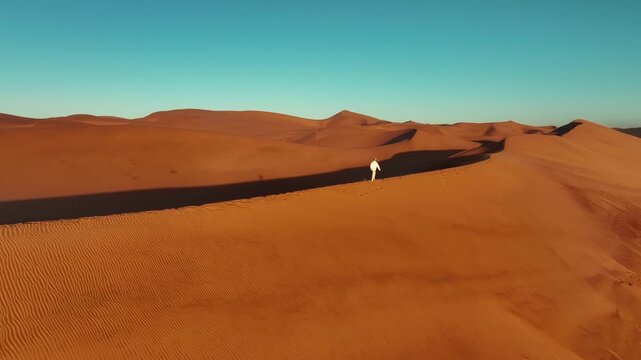 Stunning aerial drone footage of a person walking across the vast sand dunes of the Namib Desert in Namibia. Dramatic desert landscape with golden dunes stretching to the horizon