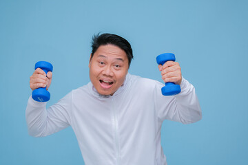 Young Asian man exercising at the gym lifting dumbbells in both hands wearing white t-shirt with happy laughing expression on turquoise blue background 