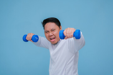 Young Asian man exercising at the gym lifting dumbbells in both hands wearing white t-shirt with happy laughing expression on turquoise blue background 
