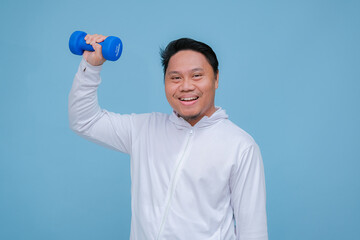 Young Asian man exercising at the gym lifting dumbbells in both hands wearing white t-shirt with happy laughing expression on turquoise blue background 
