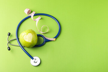 Ripe apple with carved heart, measuring tape and stethoscope on green background, flat lay. Space for text