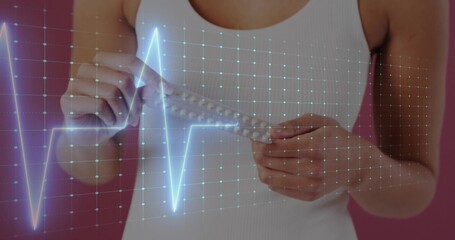 Woman holding blister pack and showing cyan ECG grid on pink backdrop, wearing white tank top